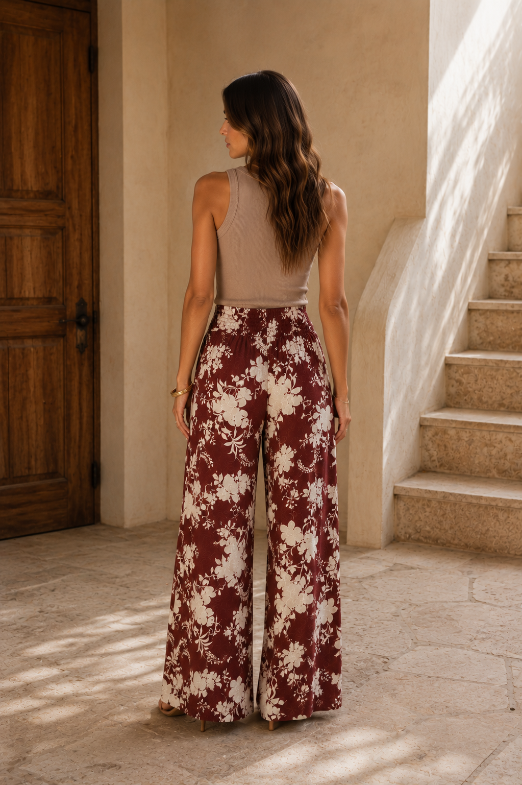 A woman stands with her back to the camera wearing a sleeveless taupe top and high-waisted, wide-leg pants with a burgundy and cream floral pattern, set in a sunlit stone corridor with a wooden door and staircase.