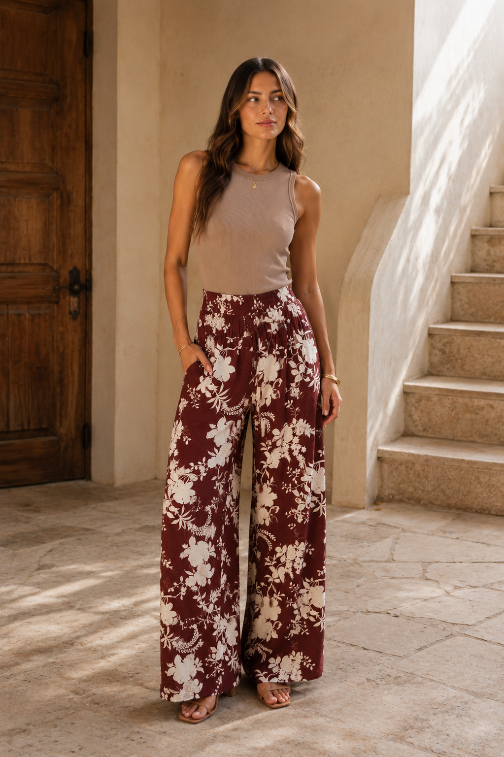 A woman stands in a sunlit architectural setting wearing a sleeveless taupe top and wide-leg pants with a burgundy and cream floral pattern.