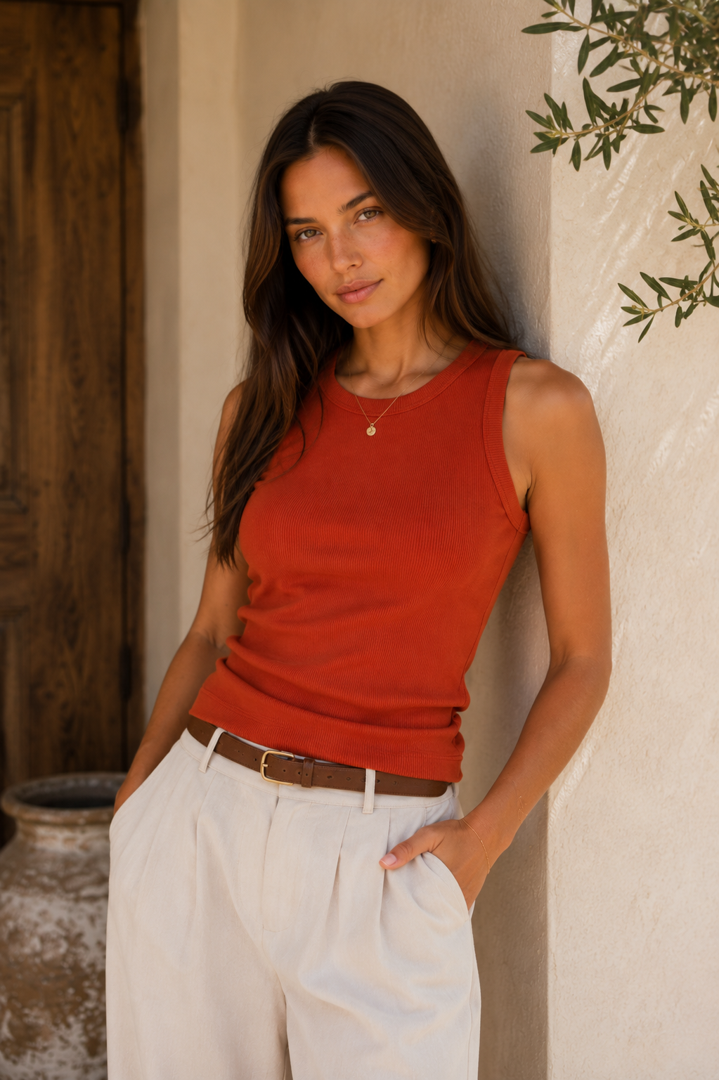 A woman wearing a sleeveless rust-orange knit top and white trousers, leaning against a textured beige wall in a warm, rustic setting.