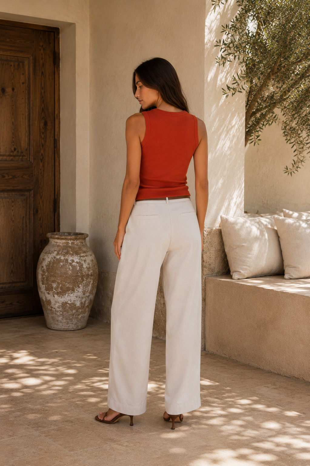A woman stands with her back to the camera in a sunlit outdoor patio setting, wearing a sleeveless rust-red top and white wide-leg pants.