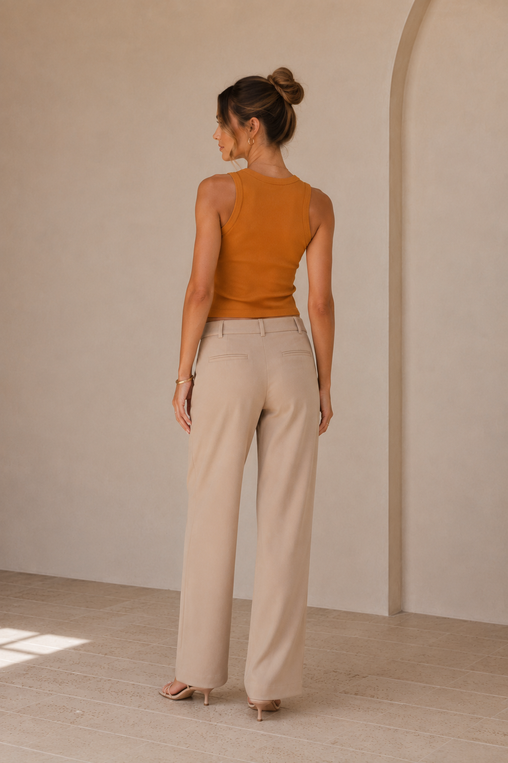 A woman shown from the back wearing a sleeveless mustard-brown top and light beige wide-leg trousers, standing in a minimalist beige interior with a visible archway.