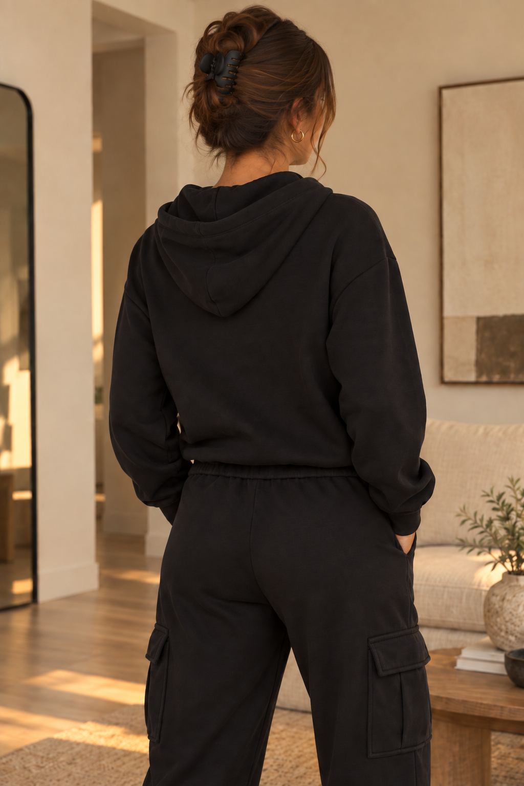 A woman viewed from behind wearing a black pullover hoodie and matching black cargo pants in a warm, modern living room setting.