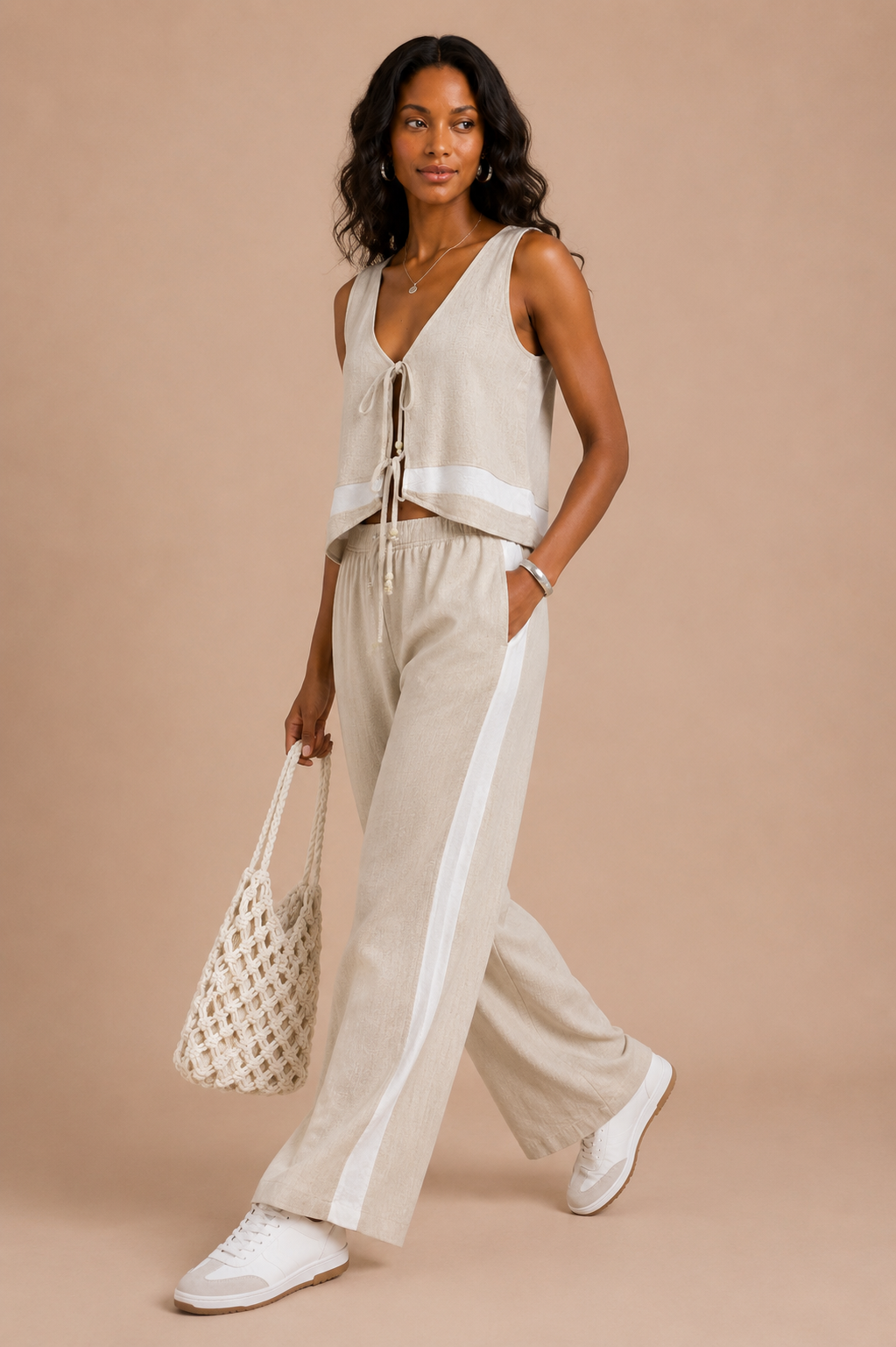 Woman wearing a beige sleeveless top and pants with white sneakers, holding a woven bag on a beige background.