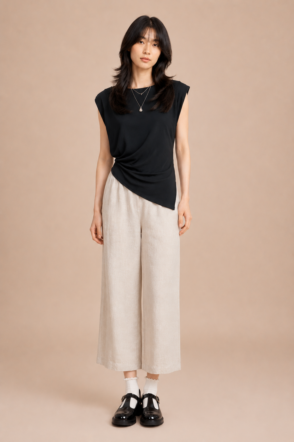 Woman wearing black asymmetrical top and linen pants against beige backdrop.