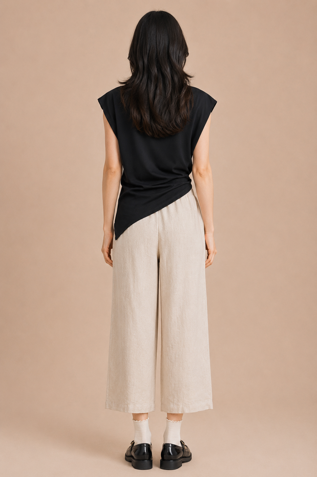 Woman wearing black asymmetrical top and linen pants against beige backdrop.