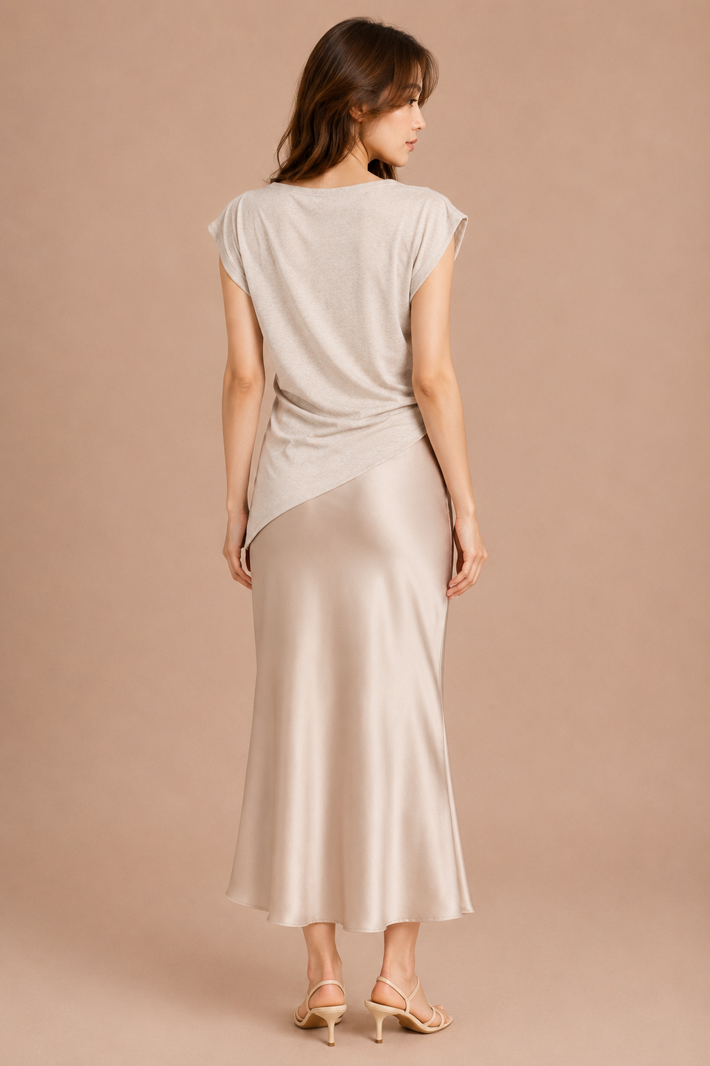 Woman wearing beige asymmetrical top and silk skirt against beige backdrop.