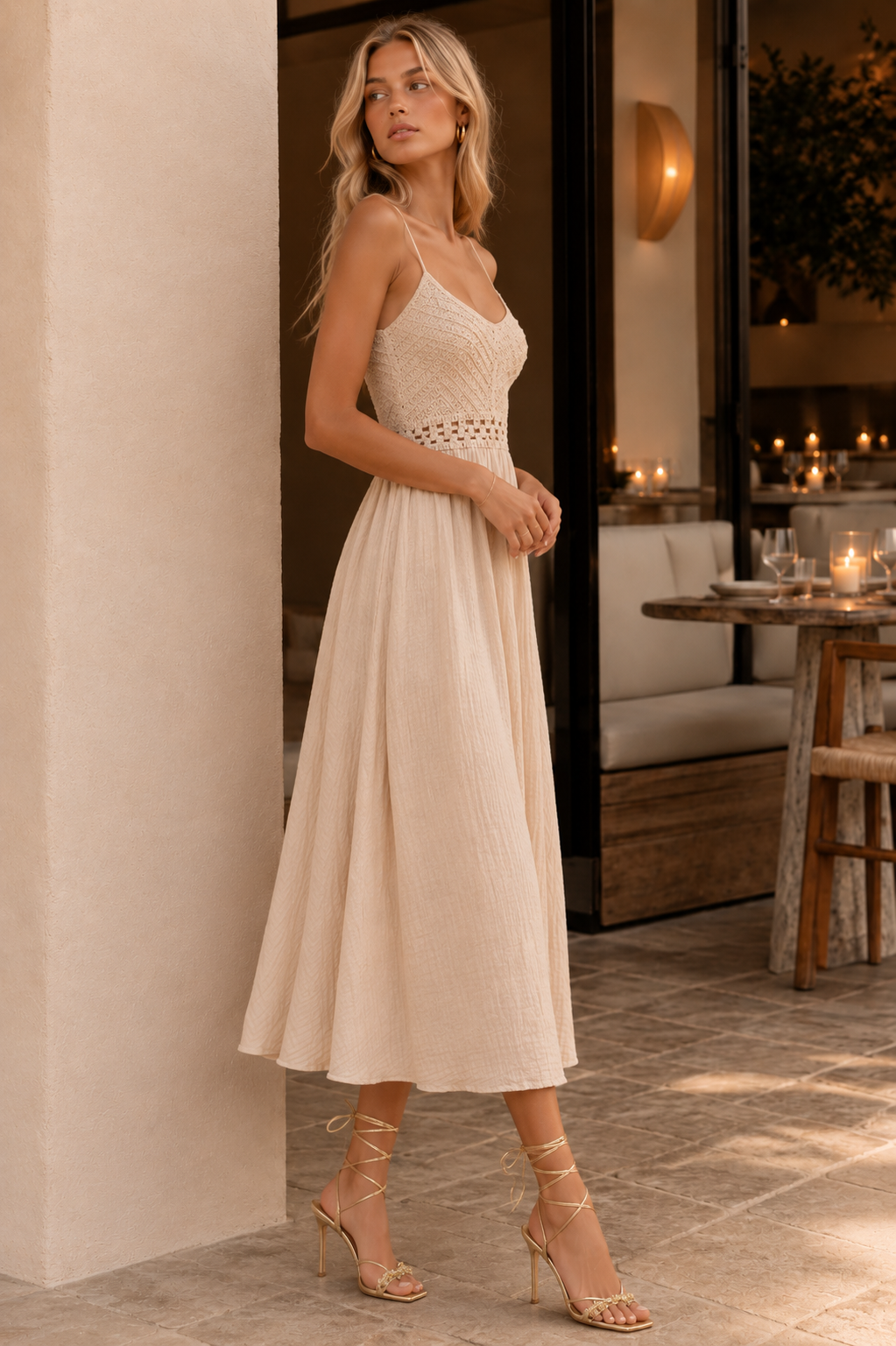A woman models a sleeveless cream midi dress with a textured lace bodice and a flowing pleated skirt, paired with nude lace-up high heels in a warm, sunlit indoor-outdoor setting.