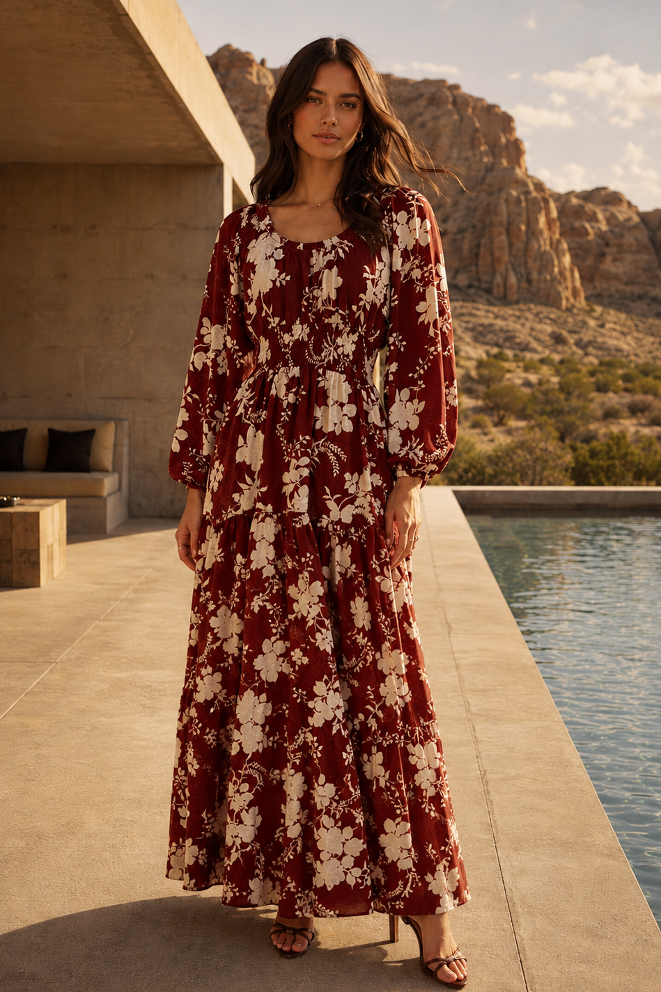 A woman stands by a pool wearing a long-sleeve maxi dress with a rust red and cream floral pattern, set against a desert landscape with rocky cliffs.